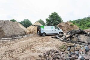 Bedrijfsbus van Ortageo met open achterklep voor 3 bergen zand. Op de voorgrond een hoop klinkers en stukken asfalt