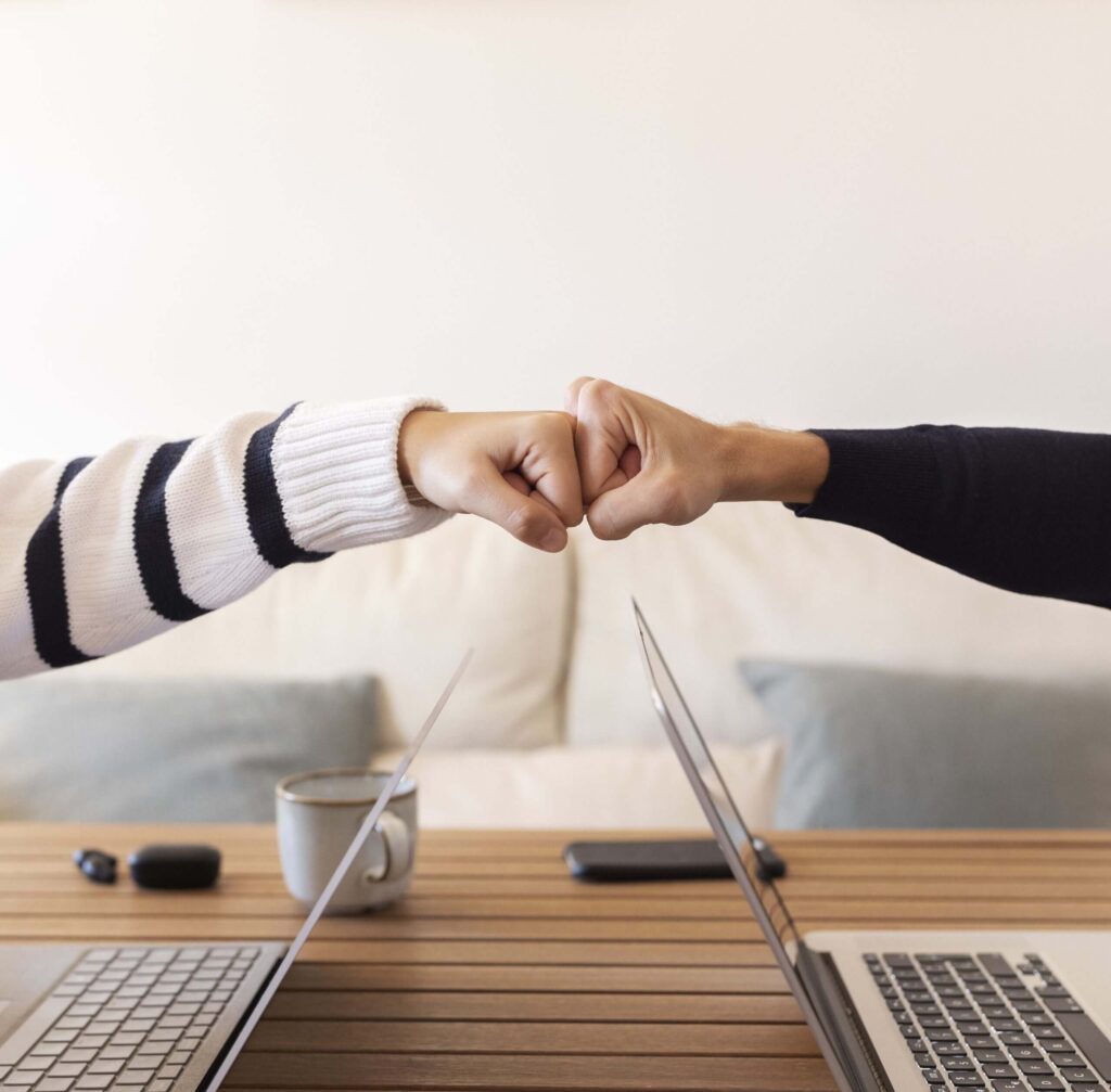 2 vuisten tegen elkaar. 1 onderarm wit met zwarte strepen trui, de andere een zwarte trui. Daaronder 2 laptops tegenover elkaar.